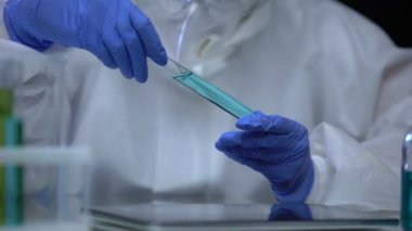 Koruyucu kıyafetli biri sıvı tutarlılığını araştırıyor, test tüpünde zehir işareti var.