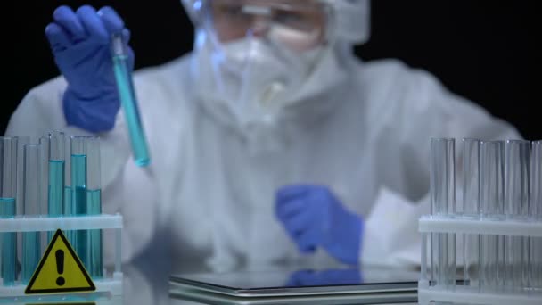 Científico en traje protector analizando tubos líquidos peligrosos, notas en la tableta — Vídeos de Stock
