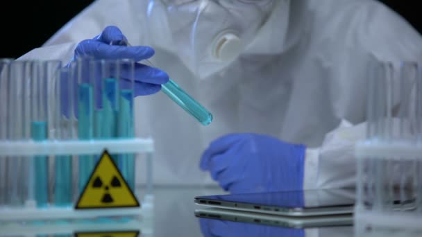 Працівник лабораторії в рукавичках тестує блакитну радіоактивну рідину в пробірці, нотатки на вкладці — стокове відео