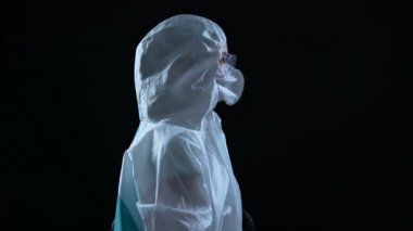 Koruyucu takım elbiseli ve maskeli laboratuvar çalışanı karanlık arka planda kameraya dönüyor.