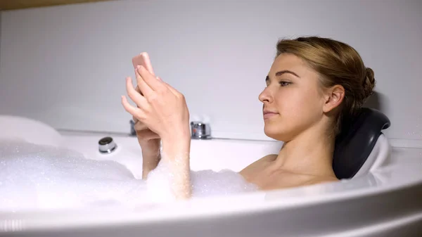 女性滚动社交媒体 躺在浴缸与泡沫泡沫 — 图库照片