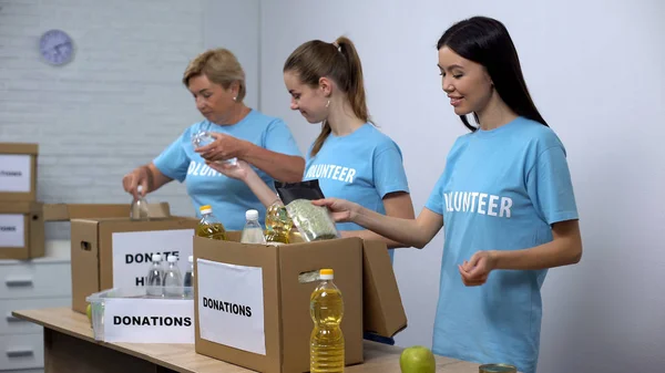 穿着志愿者T恤的欢乐妇女把食物放在盒子里 提供捐赠 — 图库照片