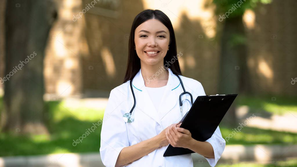 Smiling female nurse holding phone and tablet in hospital park, medical research