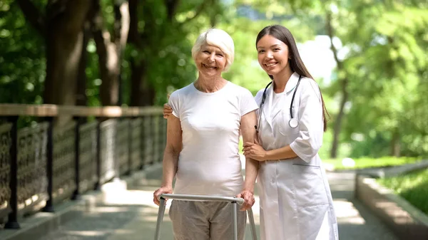 Female doctor hugging old lady with walking frame and smiling at camera outdoors