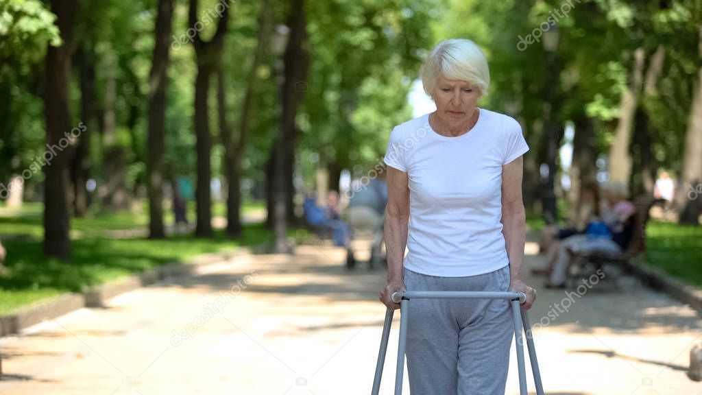 Upset old woman moving outdoors with walking frame, rehabilitation after trauma