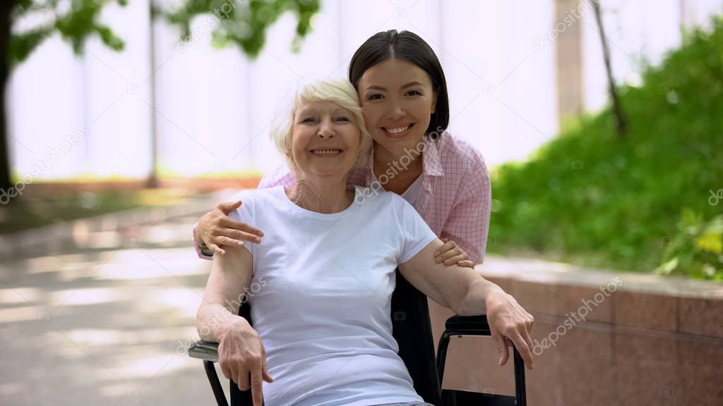 Young woman hugging grandmother in wheelchair and looking at camera outdoors