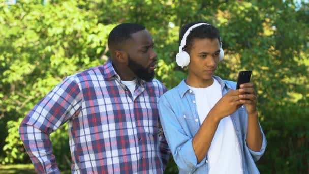 Hombre afroamericano gritando ignorando a su hijo adolescente en auriculares con teléfono inteligente — Vídeos de Stock