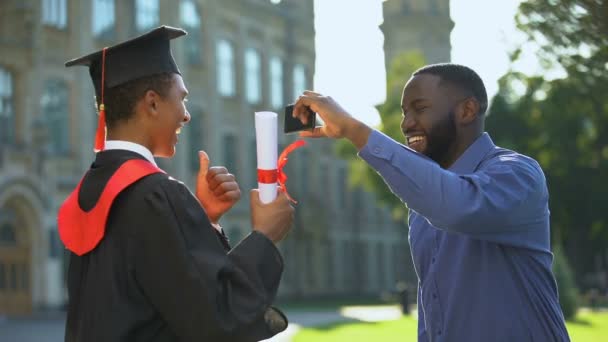 졸업 가운을 입고 십 대 아들의 사진을 찍고 있는 흑인 남자가 엄지손가락을 들고 있다 — 비디오