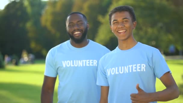 Két vidám amerikai férfi önkéntes pólóban mutogatja a hüvelykujját, önzetlen segítség. — Stock videók