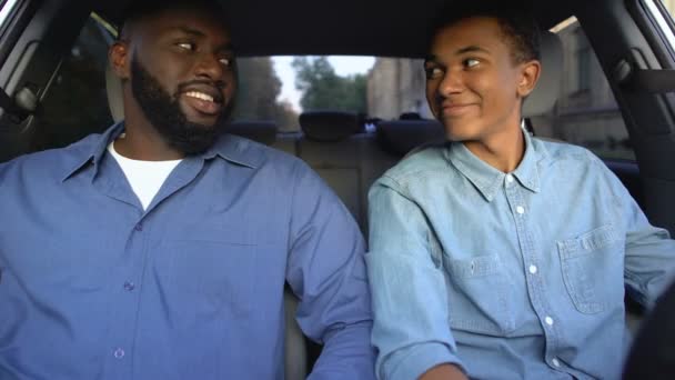 Black teenager and happy father smiling each other fastening seatbelts in car — Stock Video