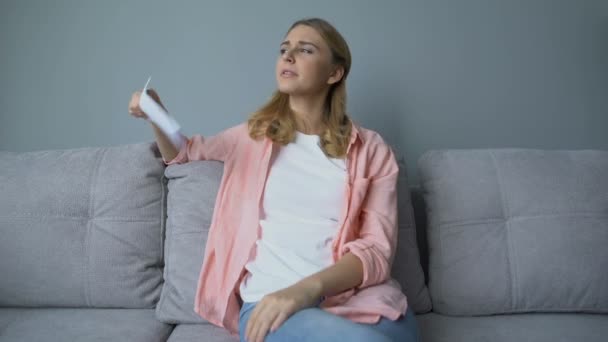Девушка машет бумагой, сидит на диване, страдает от жары, нуждается в кондиционере — стоковое видео