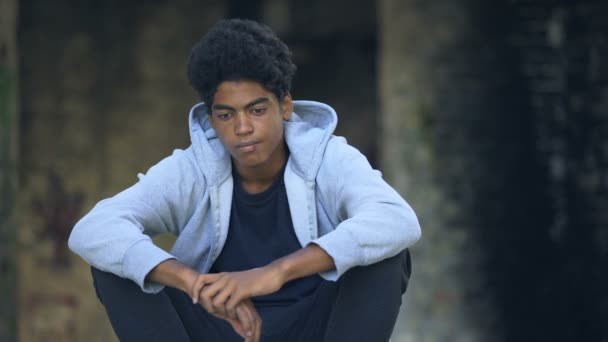 Sad afro-american teenager putting on hood sitting street alone thinking problem — ストック動画