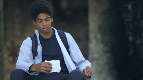 Nieszczęśliwy mężczyzna nastolatek plecak jedzenie kanapka przekąska siedzi na zewnątrz ulica życie — Wideo stockowe