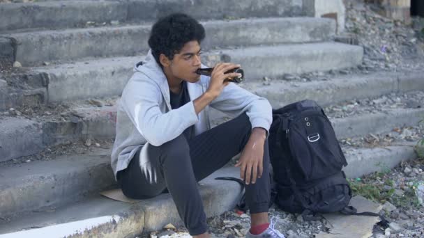 Depressed african teenager drinking beer from bottle sitting stairs, puberty age — Stock Video