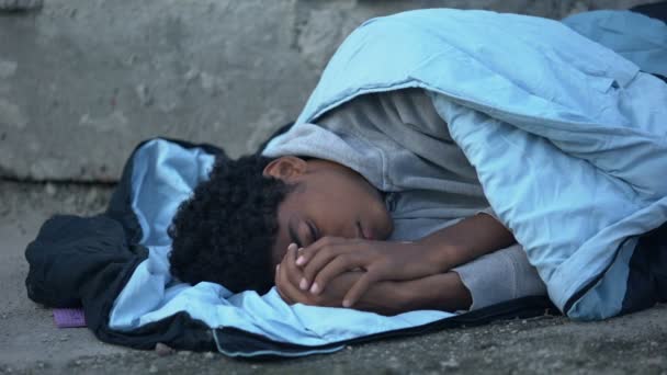 Больной чёрный подросток дрожит от холода, прикрываясь спальным мешком, лежащим на улице. — стоковое видео