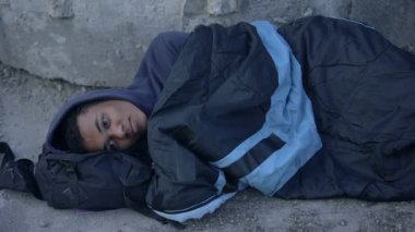 Çaresiz siyah adam uyku tulumuyla kaplı yerde yatıyor, umutsuzluk içinde.
