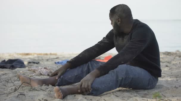 Черный мужчина плачет на берегу, бедствующий беженец выжил после кораблекрушения, катастрофы — стоковое видео