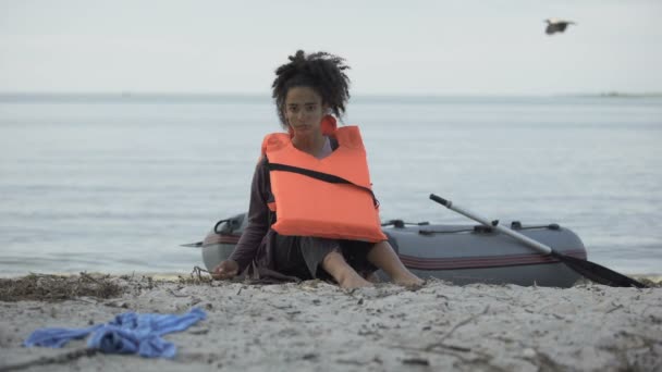 Бедная девочка-подросток в спасательном жилете сидит рядом с лодкой, беженка выжила в аварии — стоковое видео