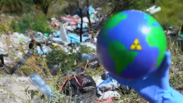 Перчатка, показывающая земной шар с радиационным знаком, свалка на заднем плане — стоковое видео