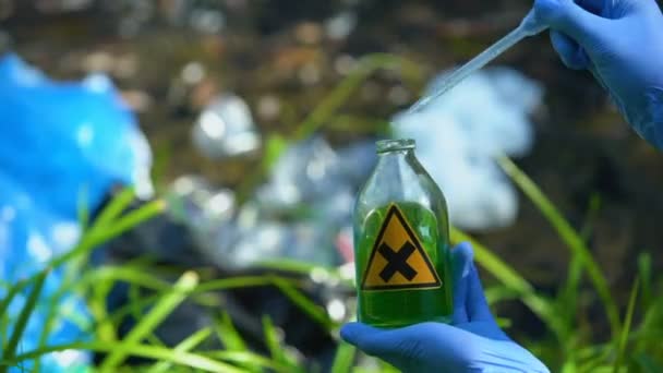 Scientist taking liquid sample from bottle with cross sign against polluted lake — ストック動画