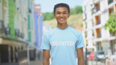 Dışarıda kameraya gülümseyen kollarını kavuşturmuş genç, melez bir erkek gönüllü.