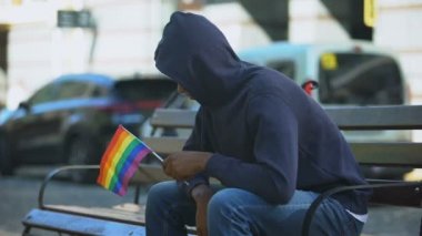 Kukuletalı siyahi adam, elinde lgbt azınlık bayrağı, önyargıları ile bankta oturuyor.