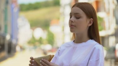 Genç kızıl saçlı kadın, şehir sokaklarında lezzetli bir sandviçin tadını çıkarıyor.