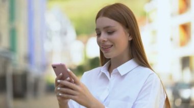 Neşeli beyaz kız dışarıda akıllı telefondan mesaj atıyor ve gülümsüyor.