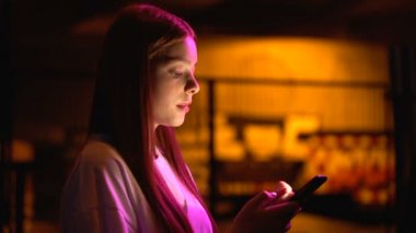 Young woman scrolling web-pages on smartphone waiting for friends near club