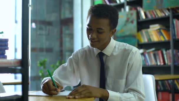 Diligente afro-americano estudiante masculino haciendo la tarea en el cuaderno, sonriendo en cam — Vídeos de Stock