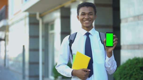 Веселий студент змішаної раси з підручниками, що показують зелений екран мобільного телефону — стокове відео