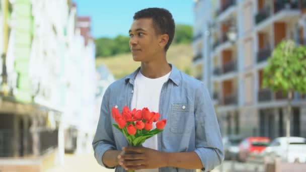 Нервный молодой мужчина с букетом тюльпанов ждет девушку на улице, первое свидание — стоковое видео
