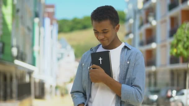 Blij Afrikaans-Amerikaanse jonge man knuffelen heilige bijbel en kijken naar kerk, geloof — Stockvideo