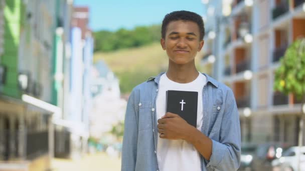 Radostný smíšený mladík držící Bibli svatou a usmívající se na kameru, náboženství — Stock video