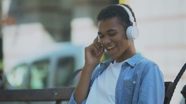 Giovane positivo che ascolta musica in cuffia e balla seduto sulla panchina — Video Stock