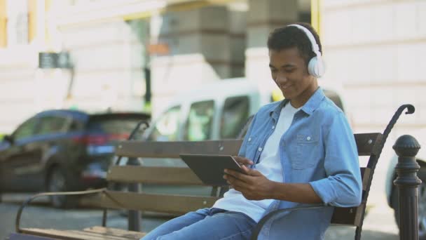 Tipo de raza mixta escuchando música en auriculares y tabletas de desplazamiento, descansando — Vídeo de stock