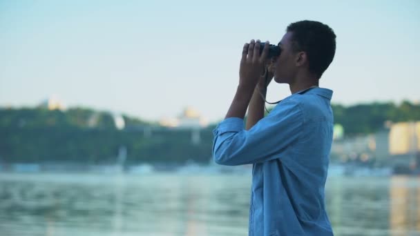 Mixed-race boy looking through binoculars at boats on river, teenager dreams — Stock Video