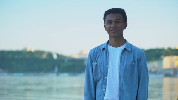 Уверенный в себе многонациональный подросток, улыбающийся стоя у реки, развлекаясь на свежем воздухе — стоковое видео