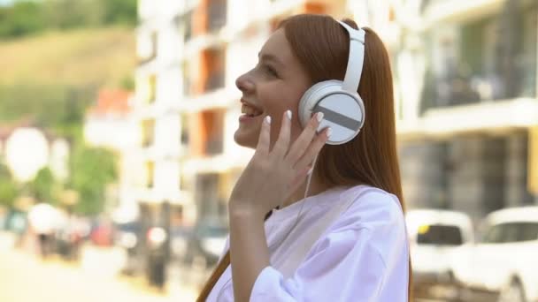 Mooie vrouwelijke tiener in koptelefoon luisteren naar favoriete muziek op straat — Stockvideo