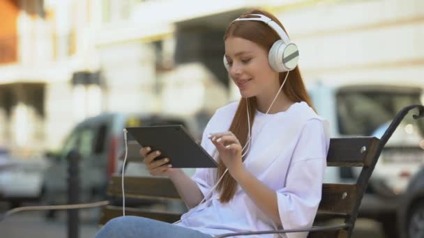 Chica pelirroja feliz en auriculares escuchando música en la tableta al aire libre, tiempo de descanso — Vídeo de stock