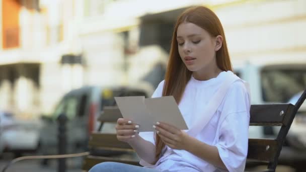 Sad red-haired girl trying to stacks pieces of torn photography sitting on bench — Stock Video