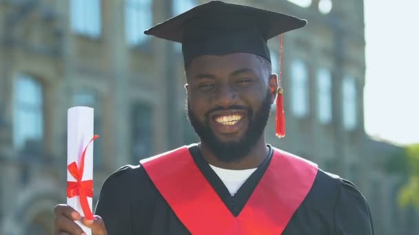 Веселий чорний студент у сукні тримає диплом і робить так жест — стокове відео