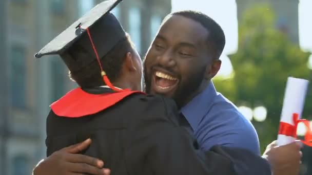 Чёрные отец и сын обнимаются в университетском городке после церемонии вручения дипломов — стоковое видео