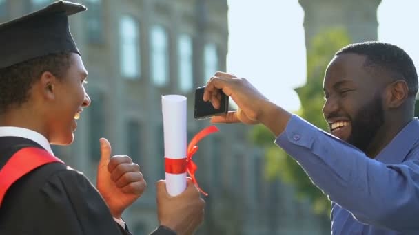 Радостный афро-американец фотографирует сына в магистратском костюме с дипломом — стоковое видео