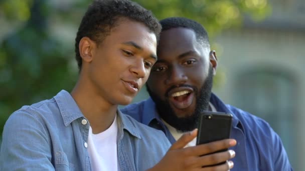 Adolescente afroamericano mostrando fotos en un teléfono inteligente a un hombre sonriente, familia — Vídeos de Stock