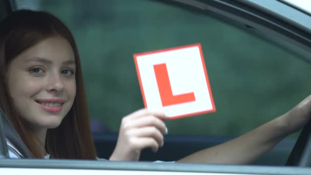 Happy student showing L-plate sitting auto, driving test, red-haired female car — Stock Video