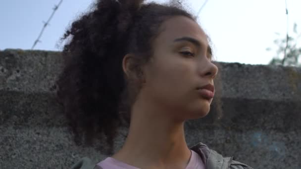 Upset African-American girl with bag standing behind wall with barber wire — Stock Video