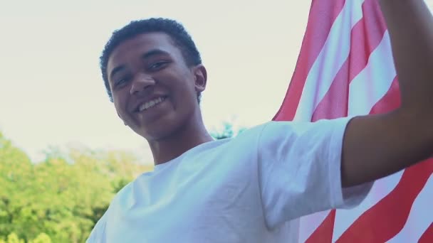 Afroamericano adolescente chico con bandera de EE.UU. sonriendo a la cámara al aire libre, día de la independencia — Vídeo de stock