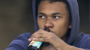 Melankolik Afro-Amerikalı, burnu yaralı, kapüşonlu genç adam şişe bira içiyor.