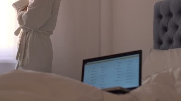 Bornozdaki kadın meyve suyu içiyor ve pencereye bakıyor, yatakta dizüstü bilgisayar, serbest — Stok video